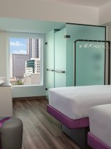 A hotel room with two single beds, each with a purple base. The room has light wood floors and an abstract purple pattern on the back wall. The bathroom is visible through a glass enclosure to the left, and a TV is mounted on the far wall with purple backlighting.