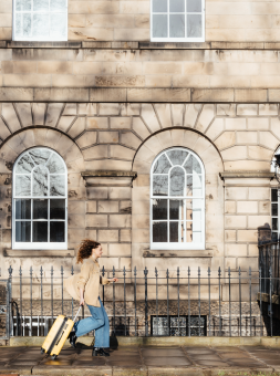 A girl pulls her suitcase along a Georgian-style street with old bricks and large pained windows. 