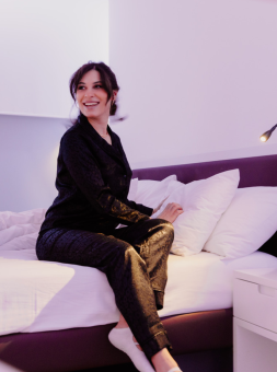 Woman dressed in smart attire sits on her hotel room bed, crossing her legs