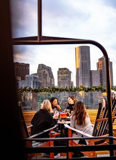 Four friends enjoy apres at a rooftop bar with Boston's skyline in the background