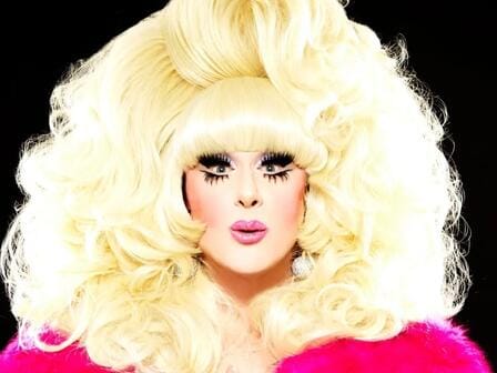 A performer in a large blonde wig and heavy makeup preparing to take the stage
