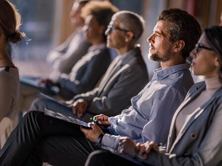 A smartly dressed audience watch an off-frame speaker intently whilst taking noes