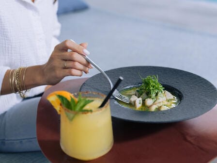 A woman in a white shirt uses a fork to eat pasta on a small table accompanied by a fruity cocktail