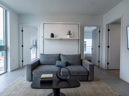 A minimalist living area with a gray couch and a black coffee table. Above the couch, a bed is folded into the wall. There are floor-to-ceiling windows on the left and a television mounted on the wall to the right.