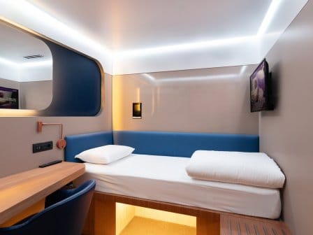 A compact, smart hotel room with a single bed, desk, mirror, TV and luggage storage space