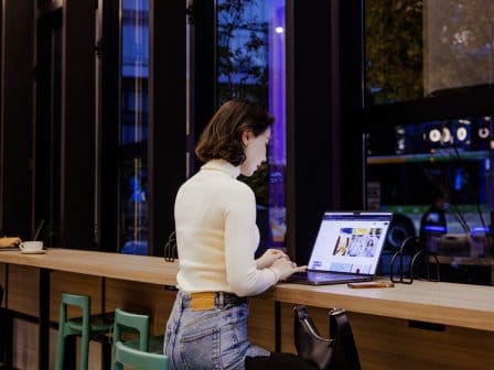 A woman browses yotel.com on her laptop in the evening while sitting by the window at a trendy restaurant and bar