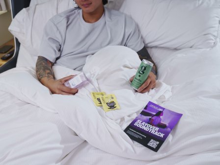 A man lies in bed holding a can of energy dink, two teabags and a flyer that reads 'Slayover Soundtrack.'