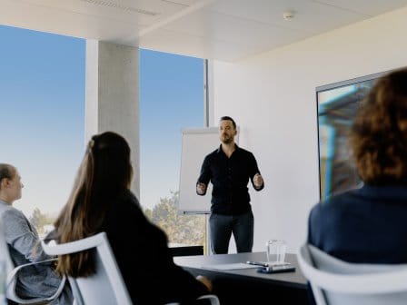 Man standing at front of room speaking to a group of people sitting at desks