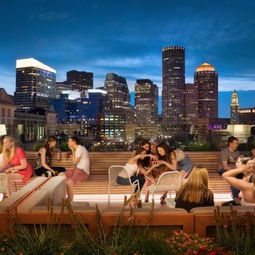 Rooftop bar at night filled with guests with the Boston skyline behind