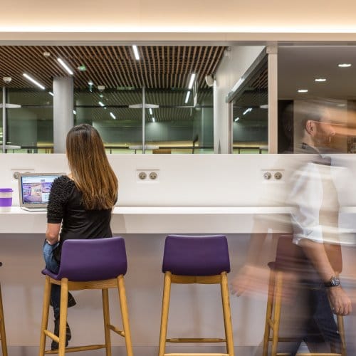 A woman in an airport hotel lounge uses her laptop as a blurry man walks by
