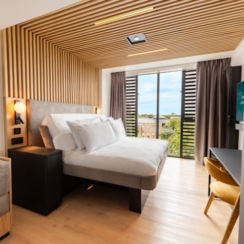 Large room with floor-to-ceiling windows, wood panelling, bedside lighting, a lounge area and a smartTV hanging from the wall.