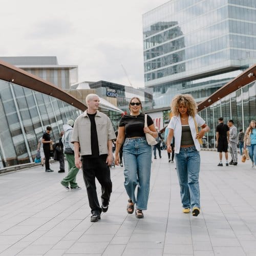 Three young adults in trendy clothes smile as they cross a modern bridge, with glistening high-rise buildings in the background