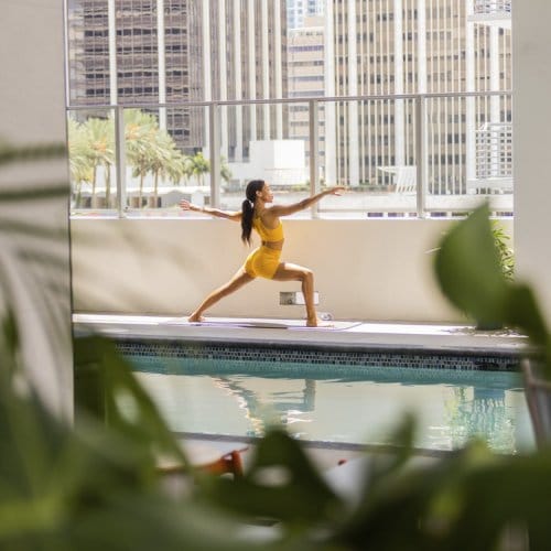 A woman practices yoga by the side of a high-rise pool with Miami Downtown skyline in the background