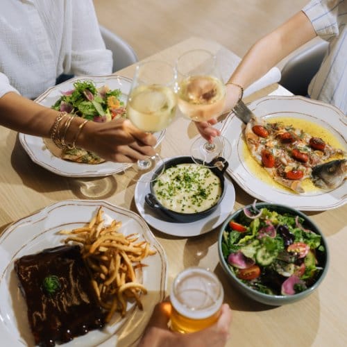 Three people clinking glasses of beer and wine over a selection of fresh food