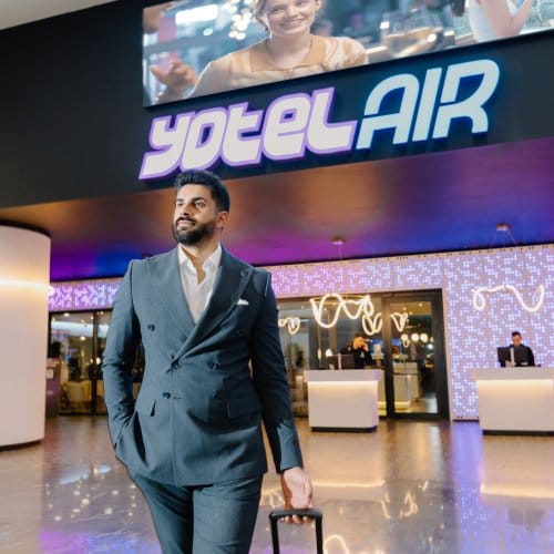 A well-dressed man rolls his suitcase outside an airport hotel facadei it reads YOTELAIR