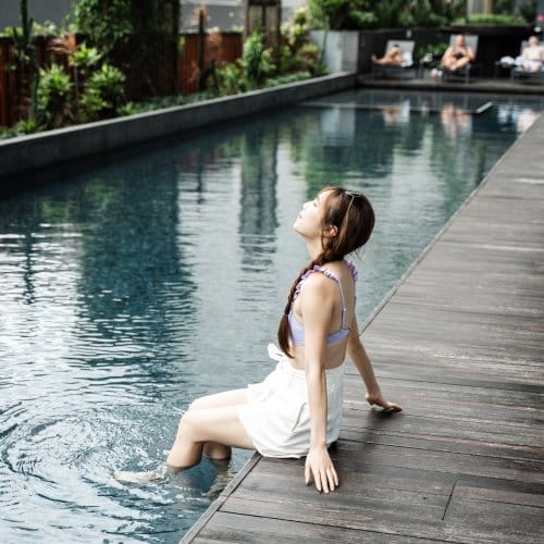 A woman dips her feet in an ourdoor hotel pool, surrounded by greenery