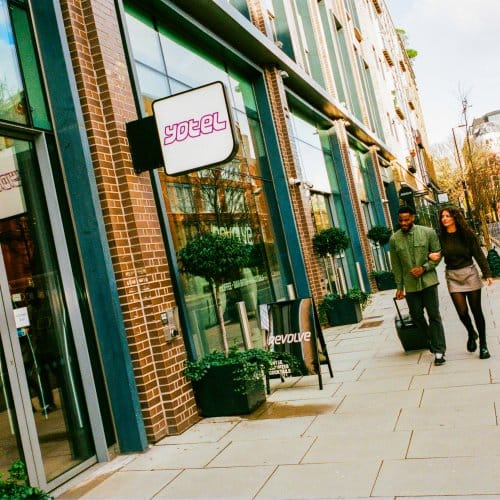 A couple walk down a sunny street approaching the front of a hotel lobby