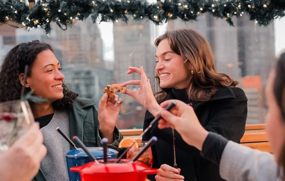 A group of friends on a cosy outdoor patio smile over hot food and cocktails