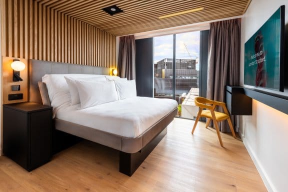 Large room with floor-to-ceiling windows, wood panelling, bedside lighting and a smartTV hanging from the wall.