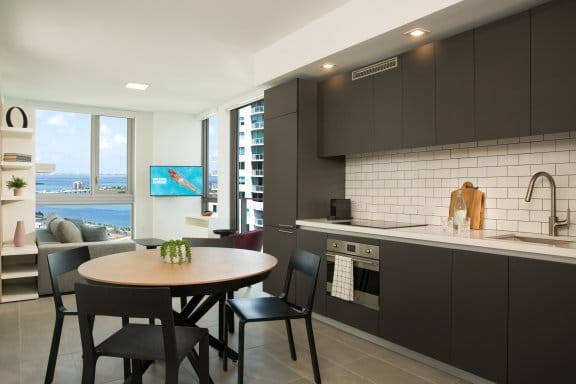 A large hotel room with generous kitchenette area, a table with chairs, huge windows and smart white-brick walls
