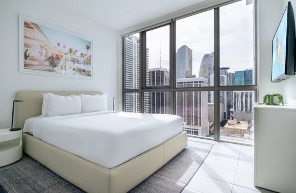 A minimalist hotel bedroom with a large bed and a framed painting on the wall. A floor-to-ceiling window offers a view of a city skyline.