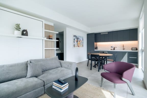 A bright, modern studio apartment with a living room and a kitchen area. A gray couch and a purple armchair face a coffee table. In the background, there's a compact kitchen with dark cabinets and a small dining table with chairs.