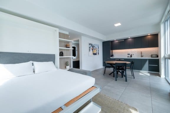 A clean and modern studio apartment. The space is divided into a bedroom area with a large bed on the left and a kitchen and dining area on the right. The kitchen has dark cabinets, and a small dining table is placed in front of it.