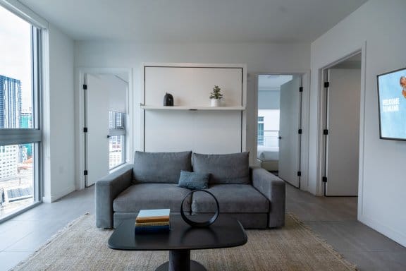 A minimalist living area with a gray couch and a black coffee table. Above the couch, a bed is folded into the wall. There are floor-to-ceiling windows on the left and a television mounted on the wall to the right.