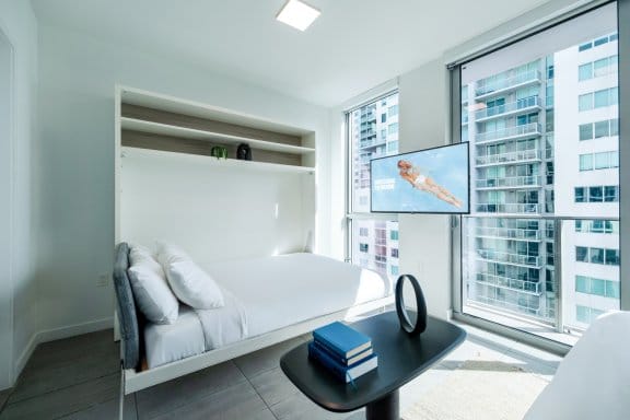 A compact and modern studio room with a bed that folds into the wall. A flat-screen TV is mounted on a stand in front of a large window with a city view, and a small coffee table is in the foreground.