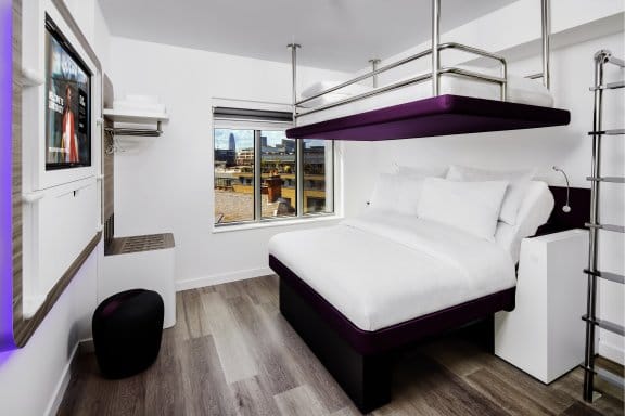  A compact hotel room with a bunk bed setup. A large bed with a purple base is on the bottom, and a twin bed with a metal railing is suspended above it. A TV is mounted on the left wall and a window overlooks a city.