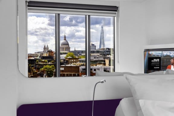 A hotel room with a large window offering a view of a city skyline. A dome, likely St. Paul's Cathedral, is prominent in the distance. A bed is partially visible in the foreground.
