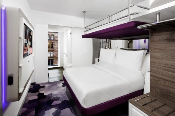  A compact, modern hotel room with a bunk bed. The lower bed is a queen size with white bedding and a purple base, and a twin bed is suspended directly above it with a metal railing. A TV is mounted on the wall to the left with purple backlighting, and a window with a view of a city street is in the background. The floor is covered in a dark, patterned rug with a geometric design.
