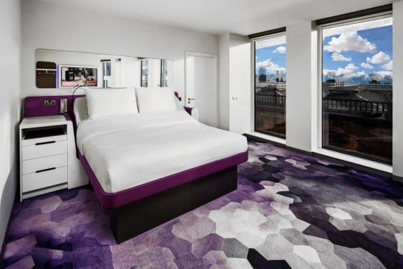 A modern hotel room with a large bed. The room has a white and purple color scheme with a dark, geometrically patterned rug. A large window offers a view of a city.