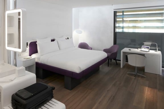A modern hotel room with a white bed featuring a purple base. The room has light wood floors, a white desk, and a large mirror with a light around it.
