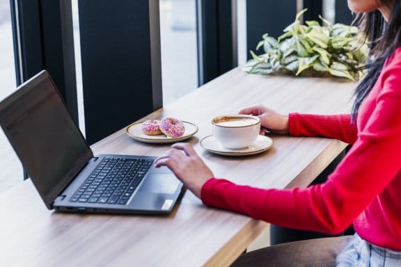 a woman works at a wooden bench on a computer, with coffee & donuts