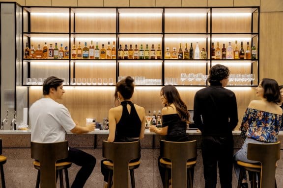 Five patrons sit in a row at a hotel bar, chatting and sipping on cocktails