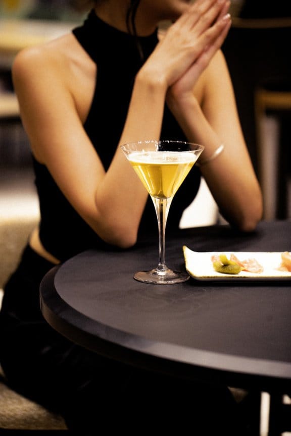 A lady clasps her hands at the table, a cocktail and canapes lie before her