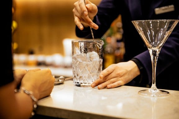 A bartender mixes a cocktail at the bar while a patron patiently waits to have a taste