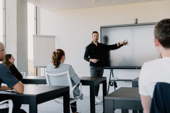 Person pointing at screen with others sitting at desks 