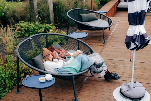 A man and a woman relax on a lounge chair snuggle on an outdoor terrace under blankets