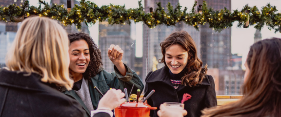 Four woman enjoy Apres on a cosy rooftop bar decorated with tinsel. The city of Boston glows in the background.