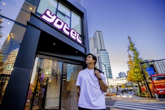 A man in a white T-Shirt holds his jacket over his shoulder while standing outside the glassy, modern exterior of YOTEL's hotel entrance