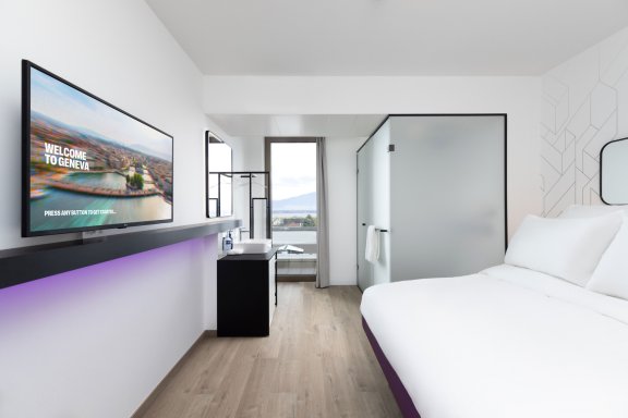 A brightly lit hotel room with white fitted sheets and a large flat-screen TV. At the end of a room, there's a large window with mountain views. 