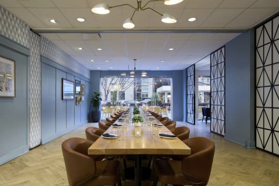 A large meeting table with street-view windows, glasses, plates and chairs set up with plants as a centerpiece