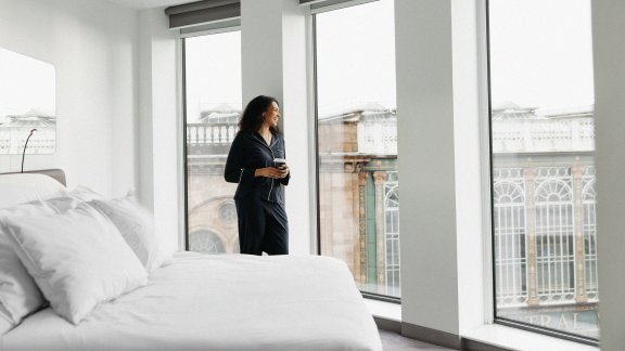A woman stands in a bright and airy hotel room with floor-to-ceiling windows overlooking a train station