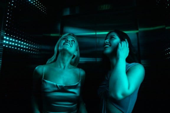 Two women stand in a lift that's flashing neon blue colours