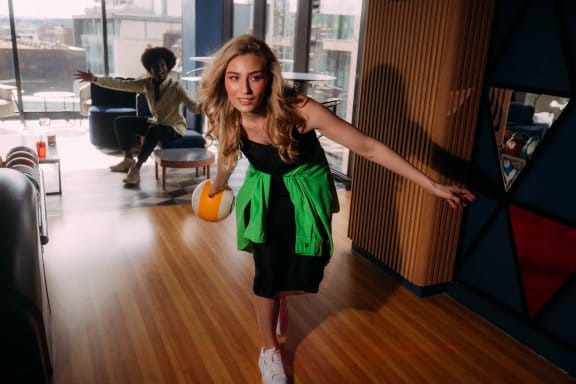 A girl rolls a bowling ball down an alley as her friend cheers on
