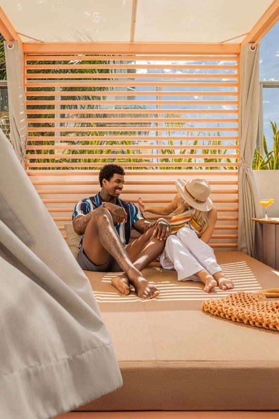 A man and a woman relax on a poolside cabana in the sun