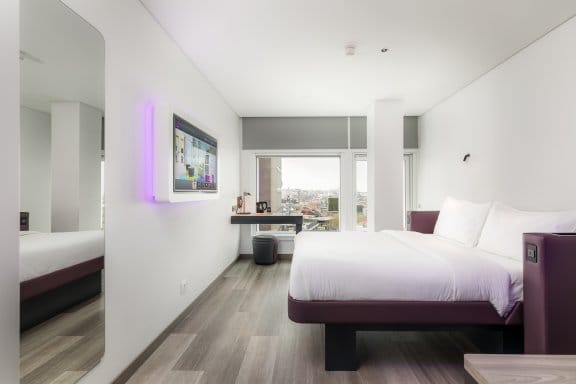 Hotel guest room depicting white and purple bed, large mirror and window