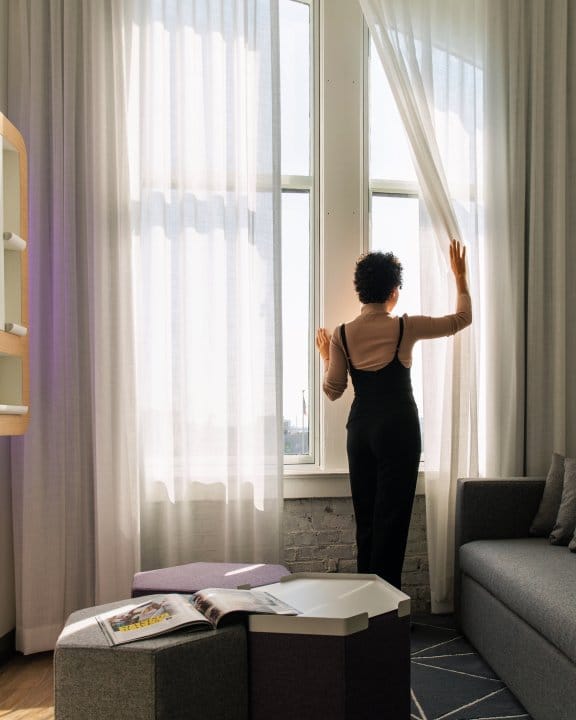 A girl in a black dress opens the curtains in her hotel suite
